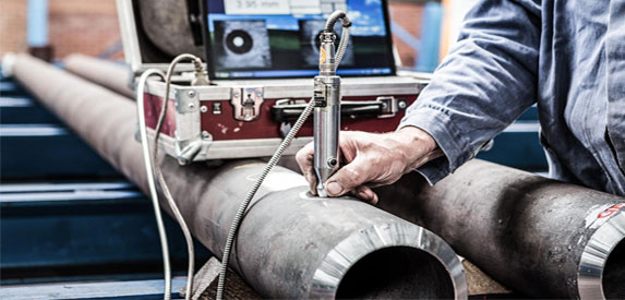 A technician inspecting metal pipes using a handheld gauge, with a digital display in the background. The scene is set in a workshop, featuring several pipes laid out on a surface, showcasing an industrial environment.
